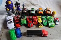 View of entire lot showing various small colorful toy cars, two purple robot-like figurines, three small humanoid character figurines, a yellow duck, and a red PEZ truck dispenser with black trailer.