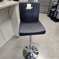 Front view of black leatherette bar stool showing square cushioned seat and backrest, chrome base and footrest.