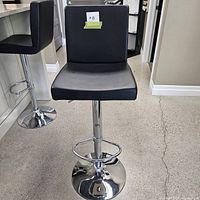 Front view of black leatherette adjustable bar stool with broad seat and backrest, showing chrome base and footrest.