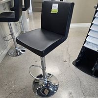 Front view of black leatherette bar stool with chrome base and footrest, showing seat, backrest, and hydraulic lift.