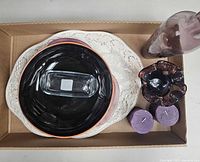 Overview of lot items in cardboard box including white turkey platter, black serving bowl, violet glassware, violet candles, and clear glass butter dish