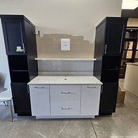 Full frontal view of two-tone vanity setup with central white drawer unit and black side pantries, quartz countertop with sink and faucet holes visible, and white tile backsplash behind.