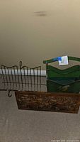 Photo showing metal wire rack with vertical wire slots and handle, wooden carved panel laid flat on floor, and green painted wood magazine rack behind them.