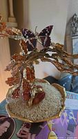 Photo showing entire bonsai tree with orange enamel leaves, metal wire branches, gold-tone tree trunk, and butterfly ornament on top, placed in a gold-tone bowl base filled with small white pebbles.