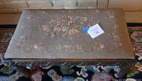 Full view of the footstool showing padded embroidered top and carved wooden frame with fluted legs.