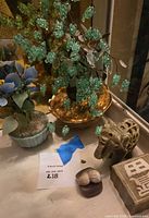 Display of decorative lot including wire tree in metallic gold bowl, ceramic flower pot with blue and yellow flowers, carved stone elephant, and marble box.