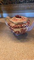 Side view of teapot showing floral and scenic red transferware pattern, handle and the hexagonal lid with 'LANG SYNE' inscription and red finial.