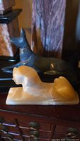Cream-colored alabaster or marble Sphinx figurine lying on a base with the blue-gray Anubis figurine in the background.