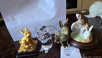 Four rabbit figurines displayed together: gold rabbit on black base, clear glass rabbit, speckled ceramic rabbit on wood base, and ivory rabbit with smaller white one on base.