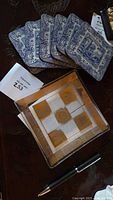 Photo of six Pumpernel corkboard coasters with blue and white scenic prints, arranged behind a square George Briard clear glass platter with gold geometric design on top of a dark surface, a pen is nearby for scale