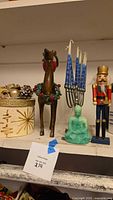Shelf display of assorted holiday figurines including wooden reindeer, green Buddha statue, metal menorah with candles, and decorative gold box with ornaments.