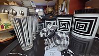 Photo showing three black and white square patterned canisters with black lids, three striped highball glasses, and a checkered rabbit figurine on a kitchen shelf.