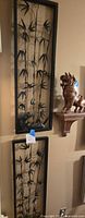 Photo showing two decorative black metal bamboo silhouette panels mounted on a beige wall, one above the other, with realistic bamboo stalks and leaves.