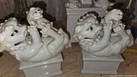 Pair of white porcelain Foo Dog figurines reclining on rectangular pedestal bases seen from above and front angles, showing details of the carved faces, raised paws, and small figures. One figurine with visible glue repair but no chips.