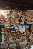 Shelf display showing various pieces of Japanese Kutani porcelain tea and coffee set including pitchers, cups, saucers, and teapots with hand-painted designs and gold accents.