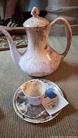 Photo of ceramic lidded pitcher with handle and spout alongside teacup and saucer featuring metallic swirl patterns