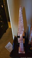 Two pink and rose marble obelisk sculptures, one taller and one shorter, shown on black bases. Visible glue repair marks on back sides of both obelisks.