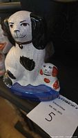 Front and side view of the white ceramic Staffordshire spaniel dog with black ears and spot, sitting with a smaller white and red puppy on a blue base. Shows hand-painted details and glazed finish with crackling.