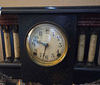 Front view of antique mantel clock showing clock face, black body with ornate engraved scrollwork, and columns on sides.