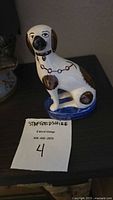Front angled photo of a ceramic Staffordshire dog figurine showing white, brown, and black painted details, blue base, and chain collar detail.