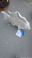 Side view of a large white ceramic fish sculpture on a concrete floor with a lot tag visible.