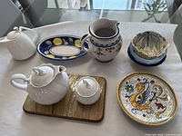 Overview of assorted servingware items including white ceramic teapot and sugar bowl on wooden tray, decorative pitcher with floral and fruit motif, blue-bordered oval dish, round peacock motif plate, and blue-rimmed covered bowls.