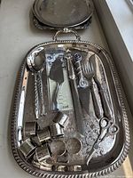 Wide view of large silver plated serving tray with assorted flatware and napkin rings arranged on it.