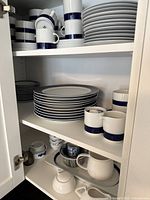 Three-shelf cabinet interior showing assorted white and blue porcelain mugs, saucers, plates and serving pieces