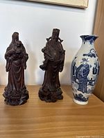 Three-piece lot showing two carved wooden Chinese statues and one blue and white porcelain vase on wooden surface.