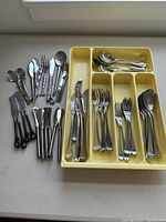 Full set of assorted stainless steel cutlery laid out including knives, forks, spoons, and specialty pieces on a table with a yellow tray.