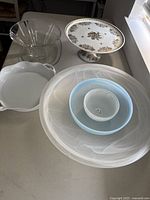 Six assorted glass and ceramic serving dishes including clear glass fluted bowl, pedestal plate with floral design, scalloped-handled dish, and three nested bowls.