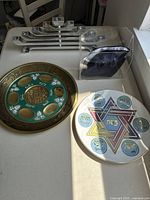 Two Seder plates side by side on a table with bright light, one gold and teal with Hebrew inscriptions, one white with Star of David and Hebrew names.