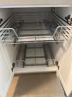 Two different views of stainless steel wire pull out kitchen cabinet organizers inside white cabinets, showing the sliding rack design with front handles and minor signs of wear.