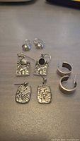 Four pairs of sterling silver earrings on a flat surface, including dome studs, wide hoops, square inset dangles, and tribal motif dangles.