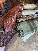 Collection of multiple vintage pillows and green plaid wool throw arranged on floor by table leg, showing intricate oriental and geometric patterned pillow designs with minor wear.