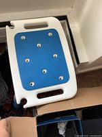 Photo of medical aid stool showing white frame with two horizontal handles, blue non-slip seating surface with drainage holes