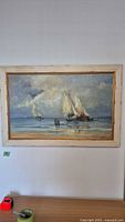 Framed original oil painting hanging on a white wall above a wooden surface, showing a seascape with sailboats and two figures standing at the shore.