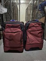 Two upright 24 inch red with black accent luggage pieces side by side, showing front zippered pockets and embroidered Roots logo.