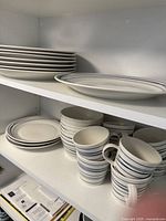 Top shelf showing stacked large white plates with thin blue stripes along edges and oval serving plates also with blue stripes.