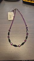 Full view of beaded necklace laid out flat, showing the alternating purple and black crystal beads and attached tag.