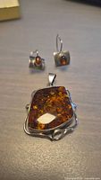 Photo showing the vintage amber earrings and pendant set in sterling silver placed on a flat surface, highlighting the pendant's large amber stone and earrings' smaller oval stones.