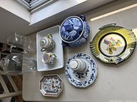 Overview of vintage ceramic items on white surface including fish-shaped plate, blue tureen, miniature teapots, octagonal dish, and stack of small plates