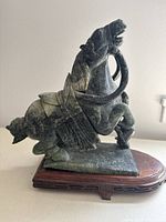 Side view of the greenish-gray carved horse statue showing detailed saddle and harness, positioned on a brown wooden base.