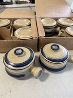 Two ceramic onion soup bowls with lids shown outside of boxes with original packaging visible in the background, highlighting the cream color with blue accents.