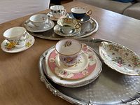 Full view of 5 teacup and saucer sets, candy dish, and silver trays showing varied floral patterns with gold trim in assorted colors on porcelain.