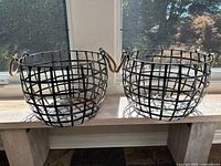 Two metal storage baskets with grid patterns and rope handles sitting on a wooden bench by a window, showing their overall size and shape.