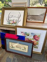 Lot of six framed art pieces arranged on a wooden shelf near a window with natural outdoor light showing varied art styles and frame finishes.