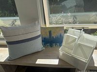 Photo showing blue and white stuffed pillow, cityscape painting, and white IKEA zip storage bins arranged on a wooden surface by a window.