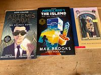 Three books visually identified: Artemis Fowl The Graphic Novel, Minecraft The Island, A Series of Unfortunate Events The Bad Beginning, placed on a wooden surface.