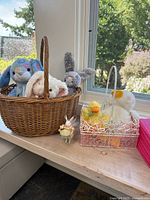 Three plush bunnies in brown woven basket, white metal basket with yellow plush chick and plush duck, decorative fairy rabbit figurine nearby on a wooden surface in front of window